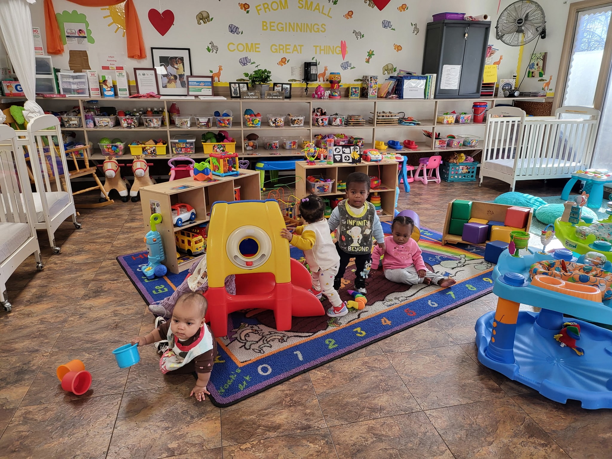 Bright infant classroom with toys, cribs, and children playing on a colorful rug