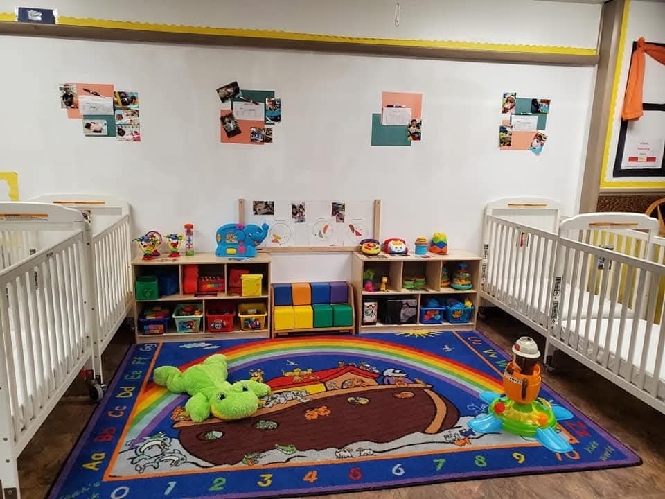Infant room with cribs, toy shelves, and a colorful Noah's Ark alphabet rug