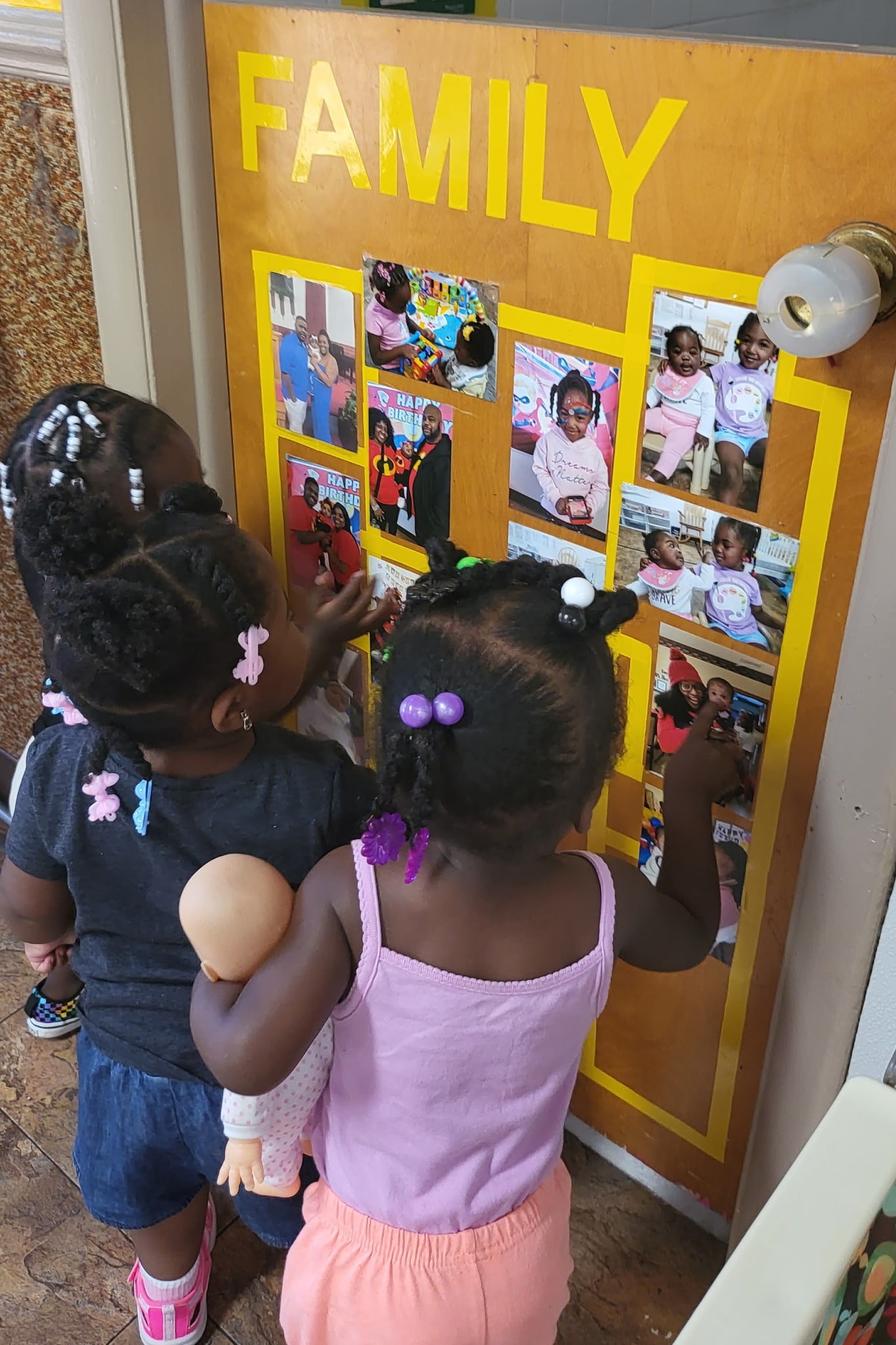 Two toddler girls looking at family photos displayed on the classroom Family board