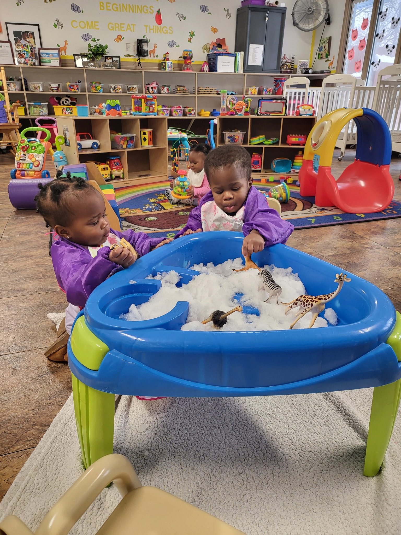 Two toddlers in purple smocks exploring a sensory bin filled with snow and animal figures