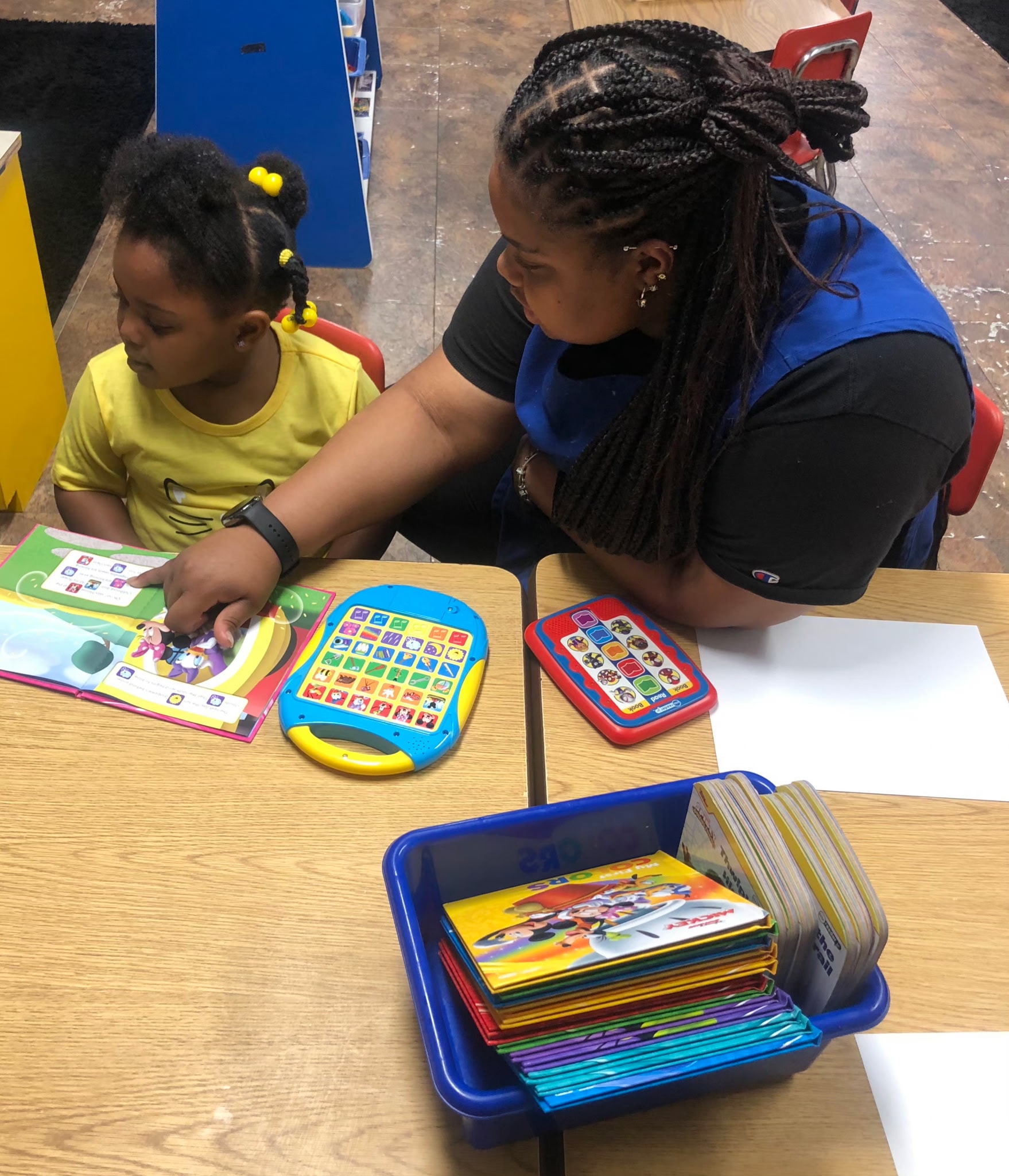 Teacher sitting with a toddler exploring interactive learning tablets and picture books at a classroom table