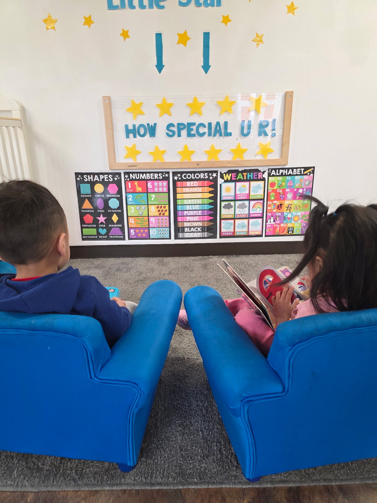 Two children sitting in blue chairs reading books in front of educational posters on shapes, numbers, colors, weather, and the alphabet