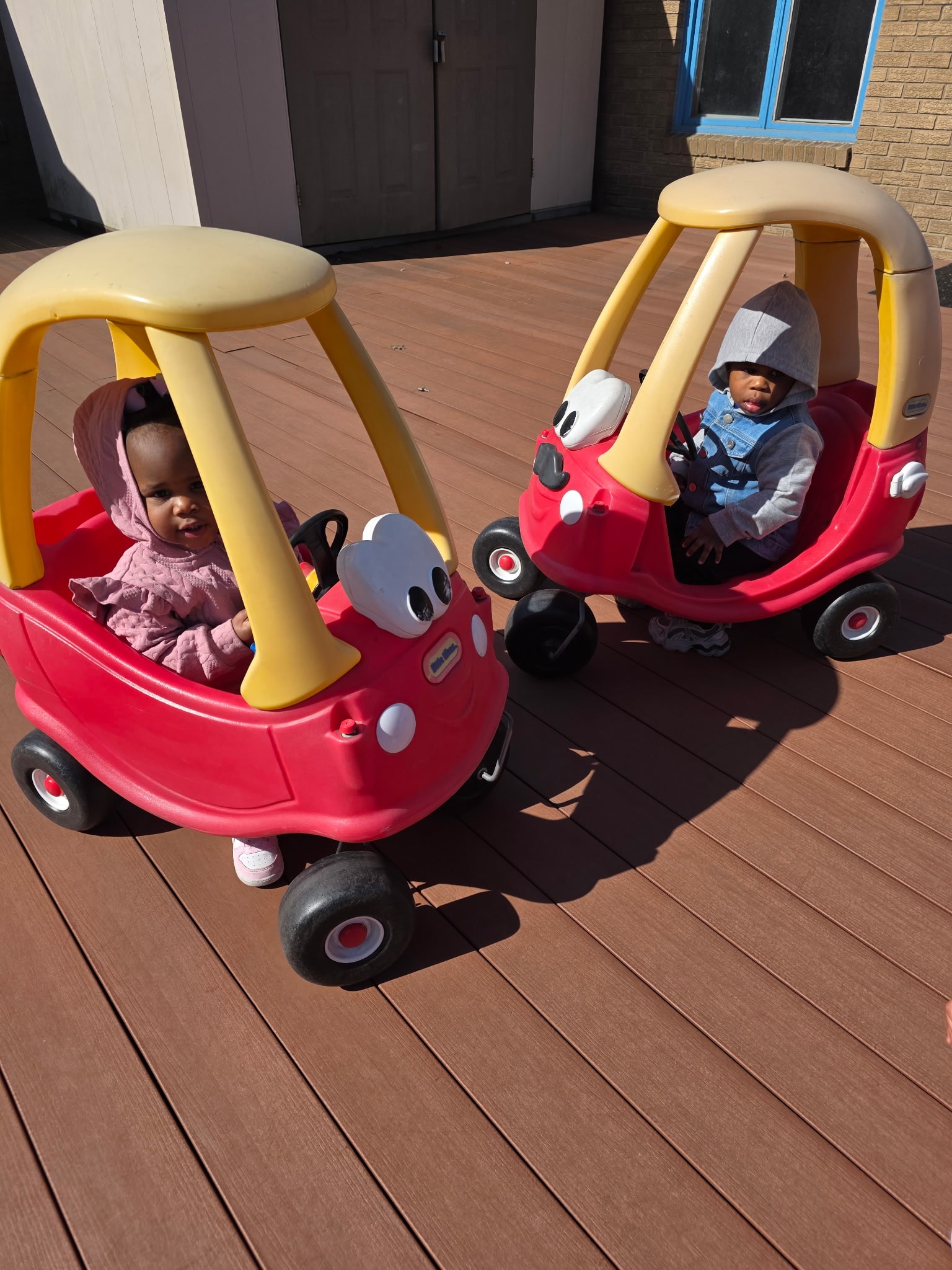 Two toddlers riding Little Tikes Cozy Coupe cars on the outdoor deck