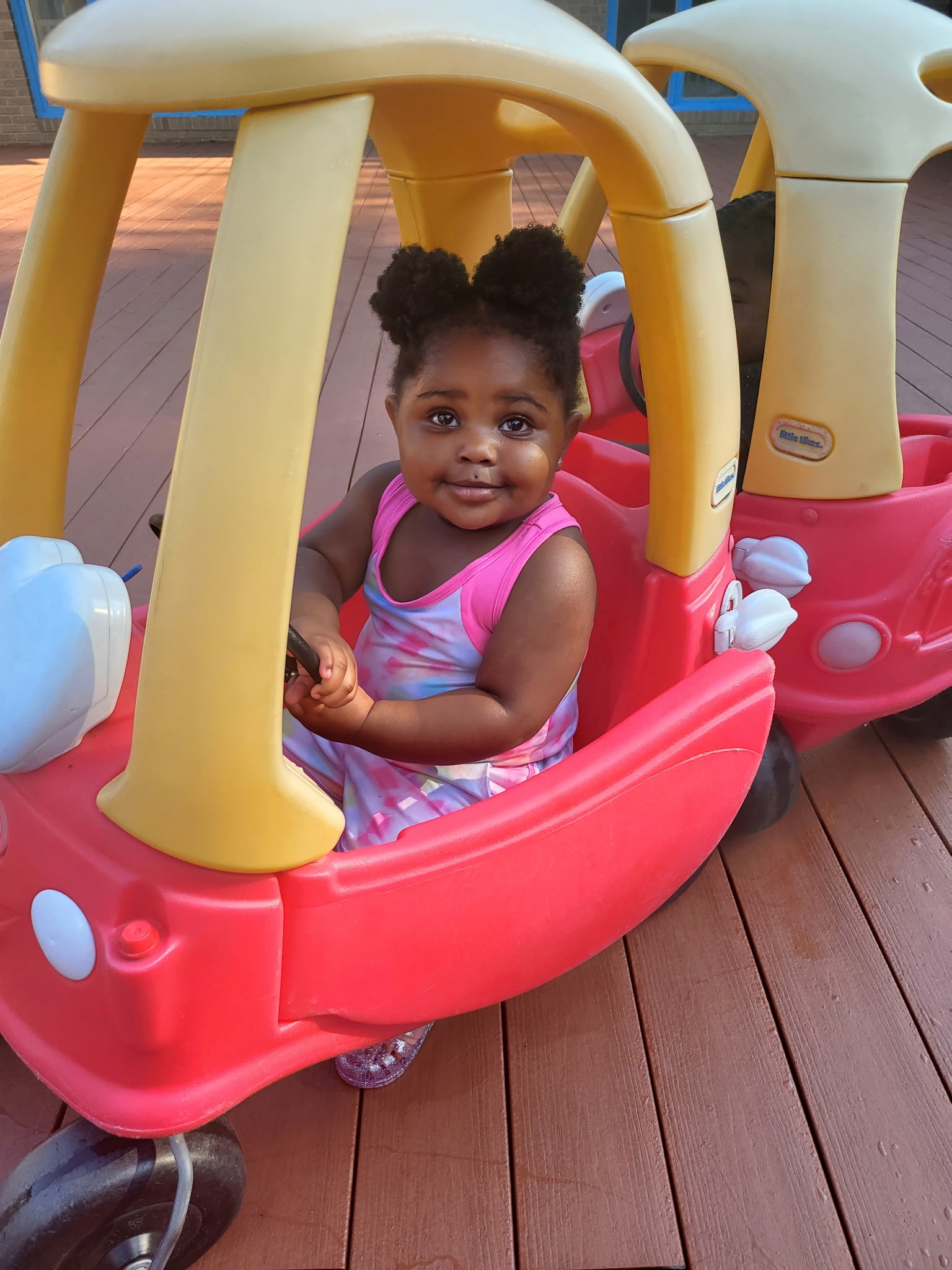 Baby girl smiling in a Little Tikes Cozy Coupe car on the outdoor deck
