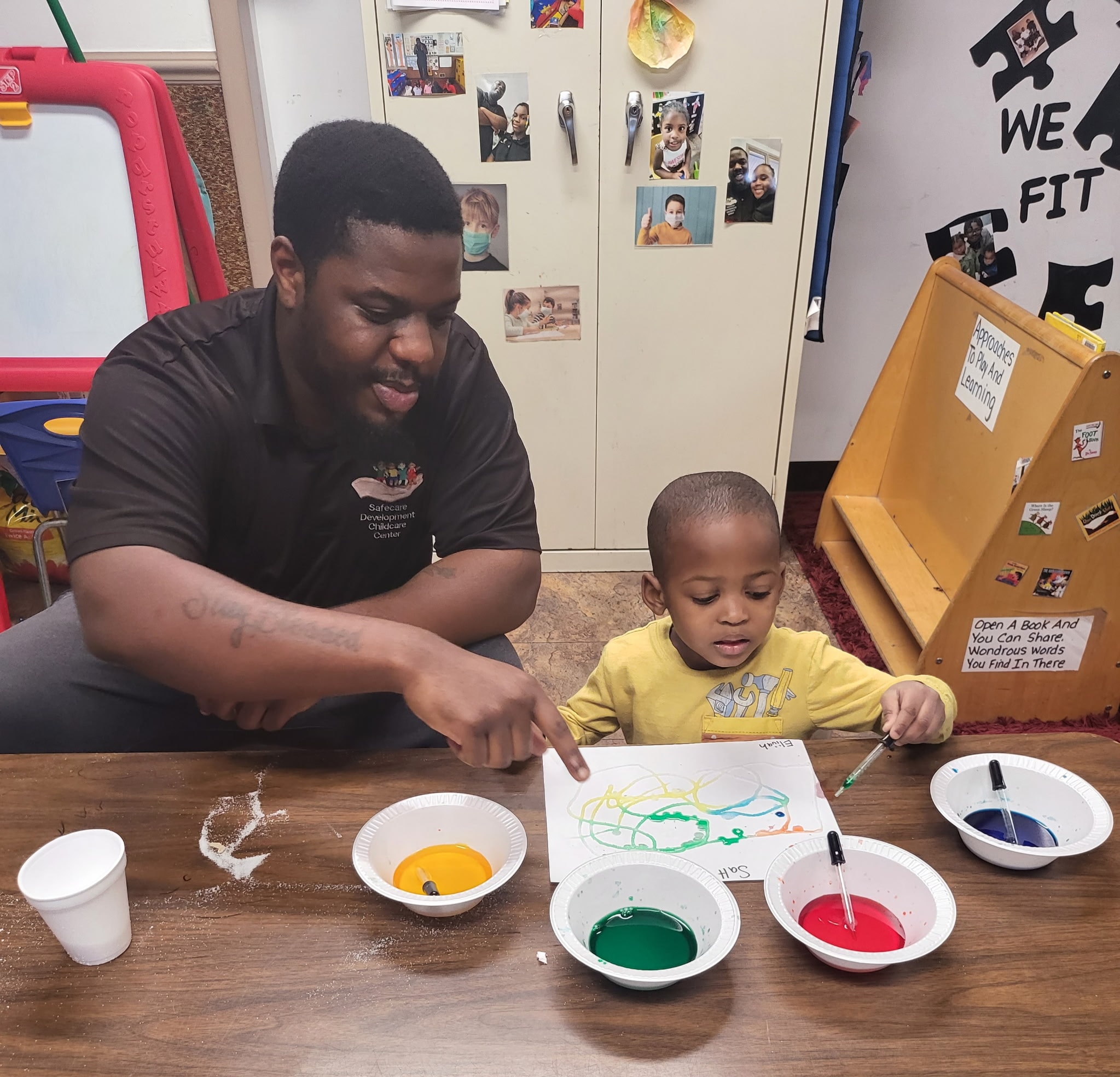 Teacher in a Safecare polo shirt sitting with a toddler doing a painting activity at a classroom table